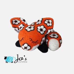 Könnte beinhalten: Ein gehäkelter orangefarbener Fuchs mit weißen und braunen Akzenten, der mit geschlossenen Augen liegt. Der Fuchs ist mit einem Granny-Square-Muster gearbeitet.