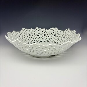 May include: A white ceramic bowl with a unique, openwork design. The bowl features a scalloped edge and is covered in a pattern of small, evenly spaced holes. The bowl is a decorative piece, perfect for displaying fruit or other items.