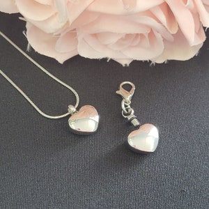 May include: Two silver heart-shaped cremation urns, one on a silver chain necklace and the other on a silver chain with a clasp. The urns are on a dark gray surface.