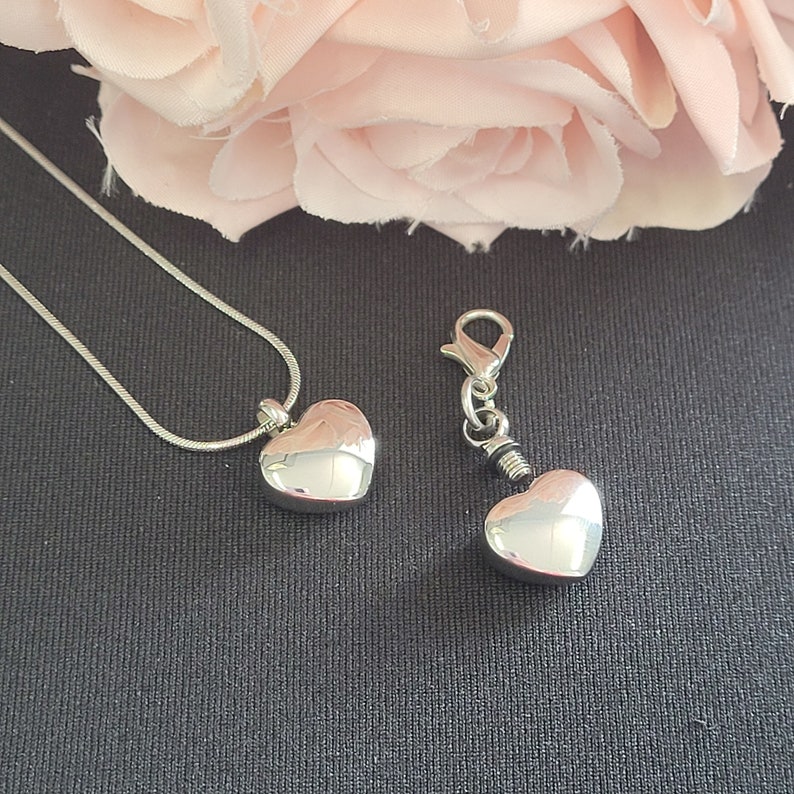 May include: Two silver heart-shaped cremation urns, one on a silver chain necklace and the other on a silver chain with a clasp. The urns are on a dark gray surface.