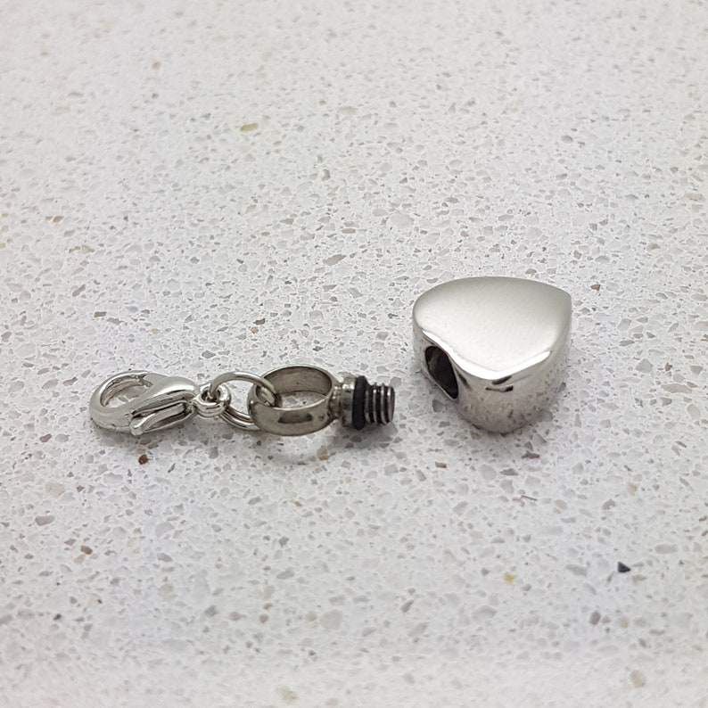 May include: A silver heart-shaped cremation pendant with a threaded opening and a separate silver connector with a clasp.