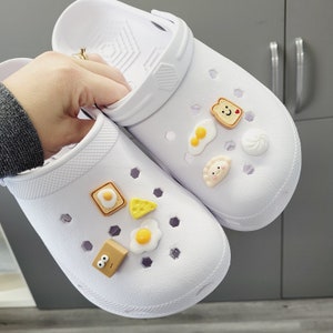 May include: Two white clogs with various food-themed charms. The charms include a fried egg, a slice of cheese, a piece of toast, a dumpling, and a swirl of whipped cream.