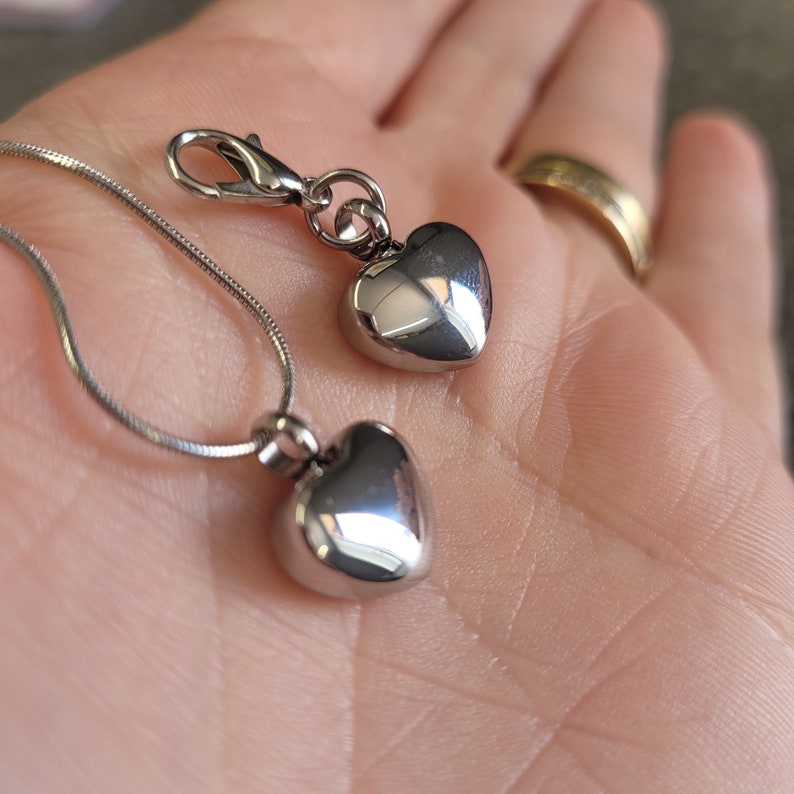May include: Two silver heart-shaped pendants on a silver chain. The larger pendant is attached to a silver clasp. The smaller pendant is attached to the chain.