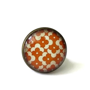 Puede incluir: Un anillo con un patrón floral redondo naranja y blanco en la parte superior. El anillo está hecho de metal con un acabado bronce.