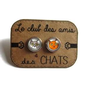 Peut inclure: Une paire de boucles d'oreilles clous sur le thème du chat sur une carte marron avec le texte "Le club des amis des CHATS". Une boucle d'oreille représente un chat gris avec des yeux jaunes et l'autre boucle d'oreille représente un chat orange avec des yeux jaunes.