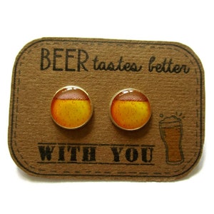 Puede incluir: Un par de pendientes de tono dorado con cabujones redondos de vidrio que representan un vaso de cerveza. Los pendientes se muestran en una tarjeta marrón con el texto "BEER tastes better WITH YOU".