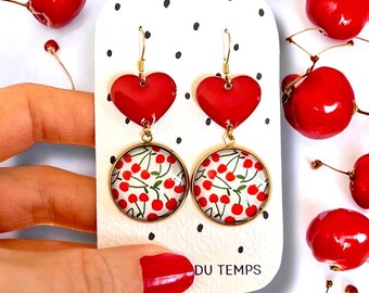 Peut inclure: Boucles d'oreilles dorées avec un cœur rouge et une base ronde ornée d'un motif de cerises. Les boucles d'oreilles sont présentées sur une carte blanche à pois noirs, avec le texte "DU TEMPS" en bas.