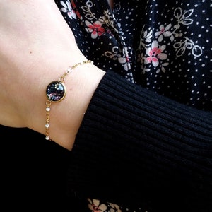 Peut inclure: Un bracelet en chaîne dorée avec un charme rond orné d'un motif floral dans les tons rose, violet et noir.