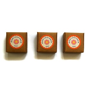 May include: Three small brown boxes with a round orange sticker on each. The sticker has a pink flower with a yellow center and the text "DANS L'AIR DU TEMPS" in a white circle with a heart at the top. The text "BIJOUX" is below the flower.
