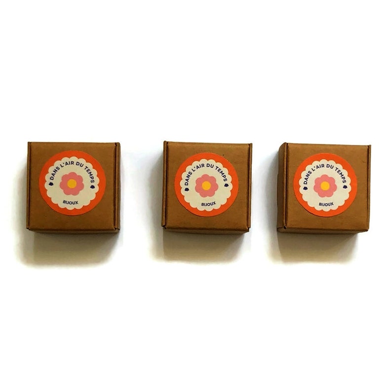 May include: Three small brown boxes with a round orange sticker on each. The sticker has a pink flower with a yellow center and the text "DANS L'AIR DU TEMPS" in a white circle with a heart at the top. The text "BIJOUX" is below the flower.