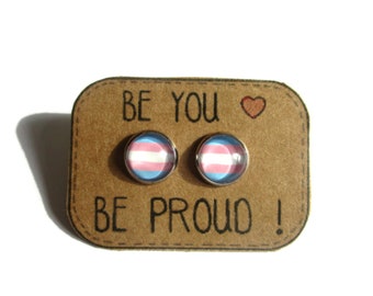 May include: A pair of stud earrings with a transgender pride flag design. The earrings are displayed on a brown card with the text "Be you" and a heart symbol, and "Be proud!"