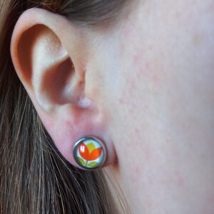 May include: A close-up of a person's ear with a small, round earring featuring a colorful flower design. The earring is silver with a red, yellow, and white flower.