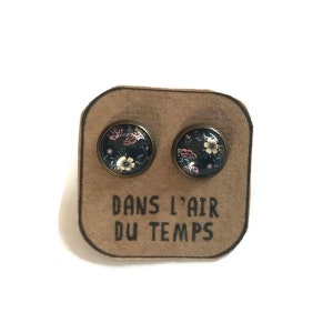 Op de afbeelding: Een paar oorbel studs met een bloemenpatroon op een zwarte achtergrond. De oorbellen worden getoond op een bruine kaart met de tekst "Dans l'air du temps".