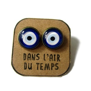 Può includere: Un paio di orecchini con l'occhio turco blu e bianco con montature dorate. Gli orecchini sono esposti su una carta marrone con il testo "Dans l'air du temps".