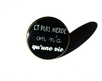 Puede incluir: Un pin redondo negro con borde dorado. El pin presenta texto blanco en cursiva que dice "Et puis merde on n'a qu'une vie". El pin está sobre un fondo blanco, con una sombra proyectada a la derecha.