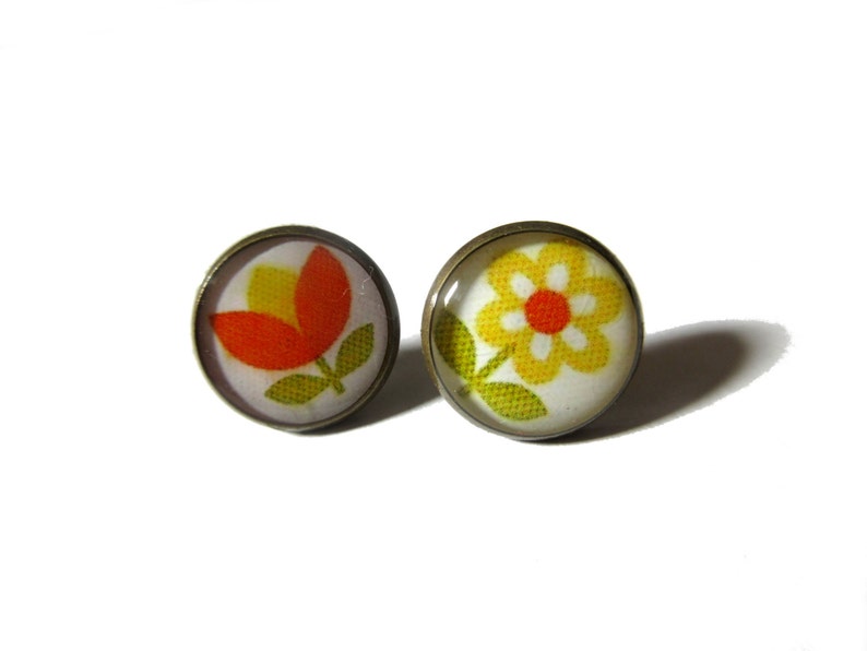 May include: A pair of earrings with a flower design. One earring features a red and yellow tulip, while the other features a yellow daisy with a red center. Both earrings have a white background and are set in a bronze-colored metal.