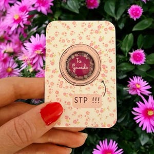 Könnte beinhalten: Eine Karte mit floralem Muster mit einem kreisförmigen Anstecker mit dem Text "Ta Gueule" in einer dekorativen Schriftart. Die Karte hat auch den Text "STP !!!" und wird vor einem Hintergrund aus rosa Blumen gehalten.