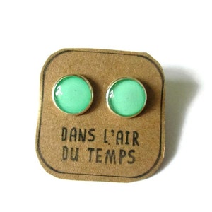 May include: A pair of small, round, mint green stud earrings with silver settings. The earrings are displayed on a brown card with the text "Dans l'air du temps".