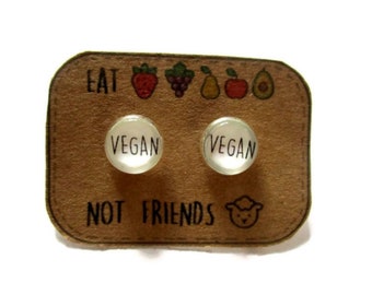 May include: A pair of small, round, white glass earrings with the word "VEGAN" printed on each. The earrings are displayed on a brown card with the text "EAT NOT FRIENDS" and a small cartoon sheep.