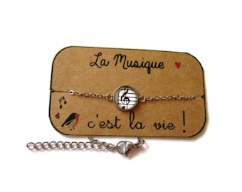 Peut inclure: Un bracelet en chaîne argentée avec un charme rond en verre représentant une clé de sol et des notes de musique sur fond blanc. Le bracelet est présenté sur une carte marron avec le texte "La Musique ♥ c'est la vie!"