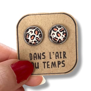 May include: A pair of round stud earrings with a leopard print design in black, peach, and white. The earrings are set in silver-colored metal and displayed on a brown card with the text "DANS L'AIR DU TEMPS".