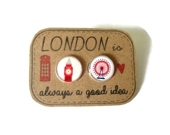 Puede incluir: Un par de pendientes con temática londinense en rojo y blanco. Un pendiente presenta un autobús de dos pisos rojo y el otro una noria roja. Los pendientes están en una tarjeta marrón con el texto "LONDON is always a good idea."