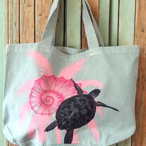 Puede incluir: Una bolsa de lona gris claro con palmeras rosas y una tortuga marina negra. La tortuga nada frente a una gran concha rosa.