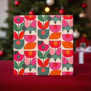 Könnte beinhalten: Eine quadratische Geschenkbox mit einem farbenfrohen Blumenmuster in Rot, Pink, Orange und Grün. Das Design zeigt stilisierte Blumen und Blätter auf cremefarbenem Hintergrund. Die Box steht auf einer roten Oberfläche mit einem Weihnachtsbaum im Hintergrund.