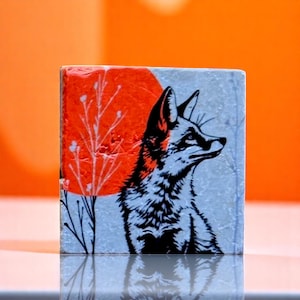 May include: Square tile with a black and white fox illustration, an orange sun, and white tree branches. The fox faces right against a light blue backdrop. The tile has a textured surface. The tile is approximately 10 cm x 10 cm.