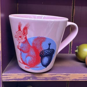 Könnte beinhalten: Hellrosa Keramiktasse mit einer Illustration eines roten Eichhörnchens und einer blauen Eichel. Die Tasse hat einen gebogenen Henkel und eine weite Öffnung. Das Eichhörnchen hält etwas in seinen Pfoten.