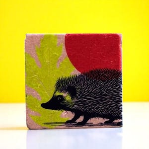 Op de afbeelding: Vierkant blok met een grillige illustratie van een egel. Het ontwerp toont een zwarte egel met stekelig haar, tegen een achtergrond van een rode cirkel en een groen bladerpatroon. Het blok heeft een getextureerd, verouderd uiterlijk.
