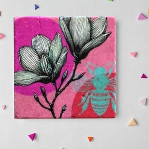 Può includere: Piastrella quadrata in ceramica con un'illustrazione botanica di fiori di magnolia in bianco e nero e una grafica di un'ape turchese su sfondo rosa. La piastrella ha un aspetto strutturato, adatta per l'arredamento.