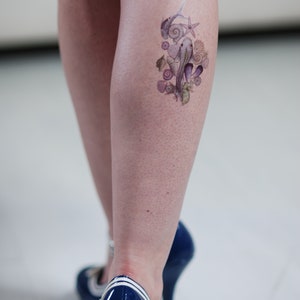 May include: A watercolour tattoo of marine life, including seahorses, seashells, and starfish, is displayed on a person's leg. The tattoo is in shades of purple, green, and beige. The person is wearing blue high heels.