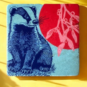 May include: Square ceramic tile with a blue and white badger illustration. A red circle with a pink mistletoe design is on the right. The tile has a light blue background, set against a yellow backdrop.