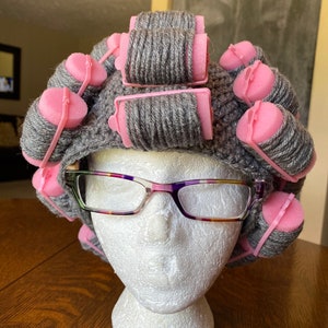 May include: A grey crocheted hat adorned with pink foam curlers and spectacles on a white mannequin head. The spectacles have a clear frame with purple, pink, and orange accents. The curlers are secured with pink plastic clips.