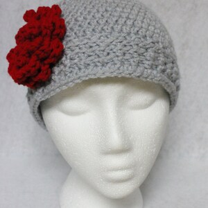 May include: A gray crocheted beanie with a prominent red flower accent. The hat is displayed on a white mannequin head, showcasing the handmade texture and design. The beanie is a stylish accessory.