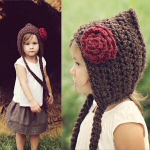 Peut inclure: Un bonnet au crochet marron avec une fleur rouge. Le bonnet a de longues tresses et est porté par un enfant. L'enfant porte un haut blanc et une jupe marron. L'arrière-plan est flou.
