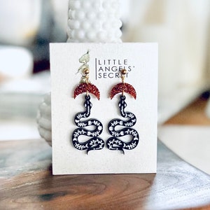 May include: A pair of black snake earrings with a red glitter crescent moon charm. The earrings are on a white card with the text "Little Angels' Secret".