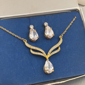 May include: A gold-toned jewelry set consisting of a necklace and a pair of earrings. The necklace features a V-shaped pendant with clear, pear-shaped stones and small pavé crystals. The matching earrings have a single pear-shaped stone.