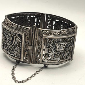 May include: Silver-toned hinged bracelet with intricate geometric and stylized figure designs. The bracelet features a rectangular clasp and a safety chain. The detailed patterns and openwork design suggest a vintage or artisan-crafted piece.