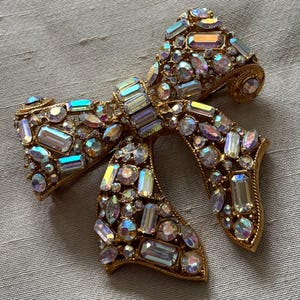 May include: A gold-toned bow brooch encrusted with iridescent, multi-shaped rhinestones. The brooch has a pin backing and is set against a neutral fabric background. The rhinestones reflect a spectrum of colors.