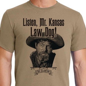 May include: A tan t-shirt with a black and white image of a man wearing a cowboy hat and the text "Listen, Mr. Kansas Law Dog!" and "Ike Clanton" in a banner.