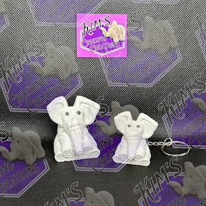 May include: Two white elephant keychains with a silver key ring. The keychains are made of a soft, textured material and have black eyes. The keychains are on a purple and black background with the text "Kim's Cruising Creations" repeated in the background.