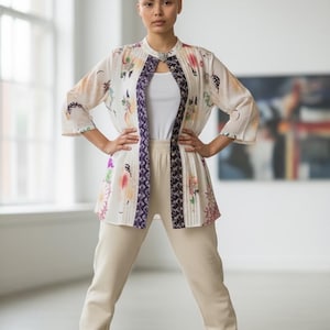 May include: A woman wearing a cream-colored outfit, including a white tank top, pants, and sneakers. The outfit is paired with a floral patterned jacket with purple trim and a decorative clasp.