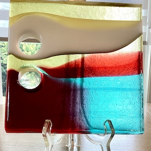 May include: A colorful glass art piece with a layered design of yellow, white, red, and blue. The piece has two circular cutouts and is displayed on a clear acrylic stand.