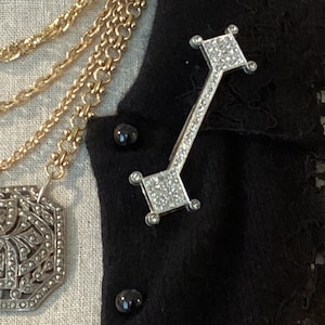 May include: A silver brooch with a geometric design, featuring a row of clear rhinestones. The brooch is pinned to a black cardigan.