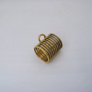 May include: A cylindrical gold-toned metal thimble featuring a series of parallel horizontal bands with a textured, etched surface and a small circular loop attached to the top edge, designed for finger protection during sewing tasks.