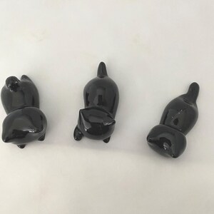 May include: Three glossy black cat ornaments in different poses. The cats have rounded bodies and small, pointed ears. The ornaments are displayed against a plain white background, highlighting their sleek, reflective surfaces. These decorative cat ornaments are ideal for cat lovers.