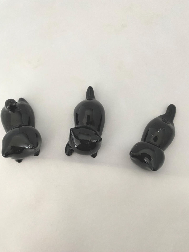 May include: Three glossy black cat ornaments in different poses. The cats have rounded bodies and small, pointed ears. The ornaments are displayed against a plain white background, highlighting their sleek, reflective surfaces. These decorative cat ornaments are ideal for cat lovers.