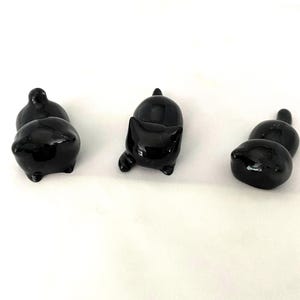 May include: Several glossy black cat ornaments in different poses. One cat is standing, another is seated, and the others are lying down. The ornaments have a smooth, reflective surface and are set against a white background.